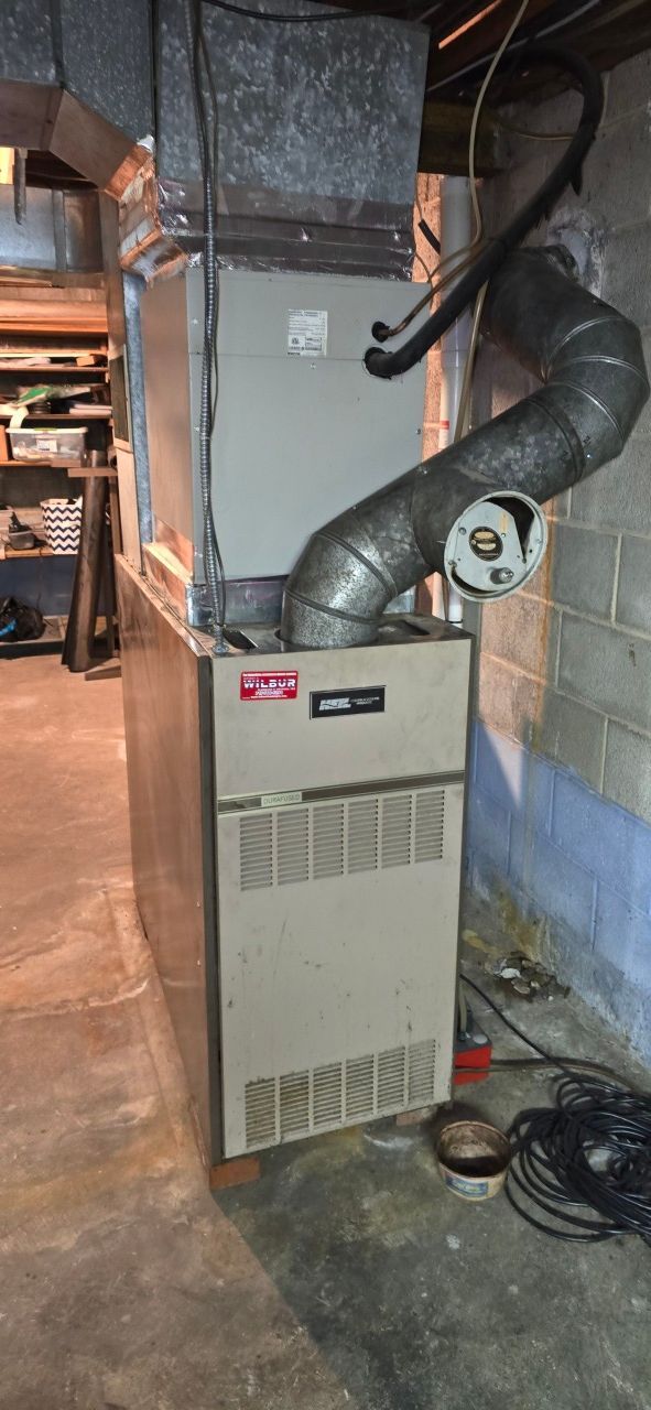 A tan HVAC furnace unit stands in a basement with metal ductwork and a curved vent pipe attached to the top.