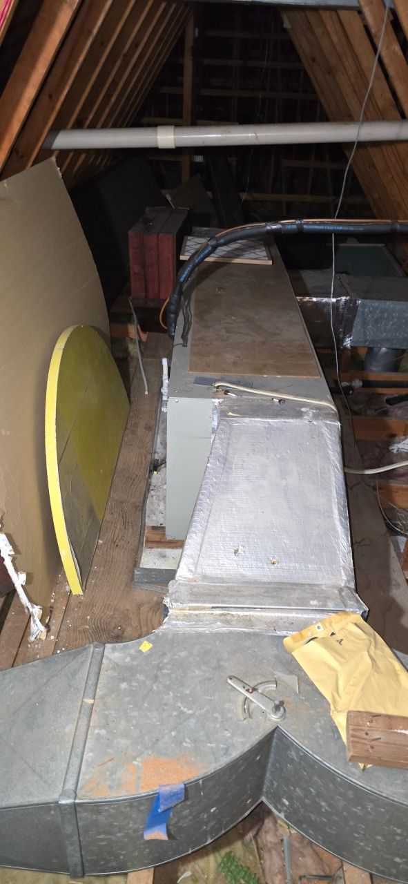 An HVAC unit and metal ductwork situated in a dimly lit, wooden-framed attic.