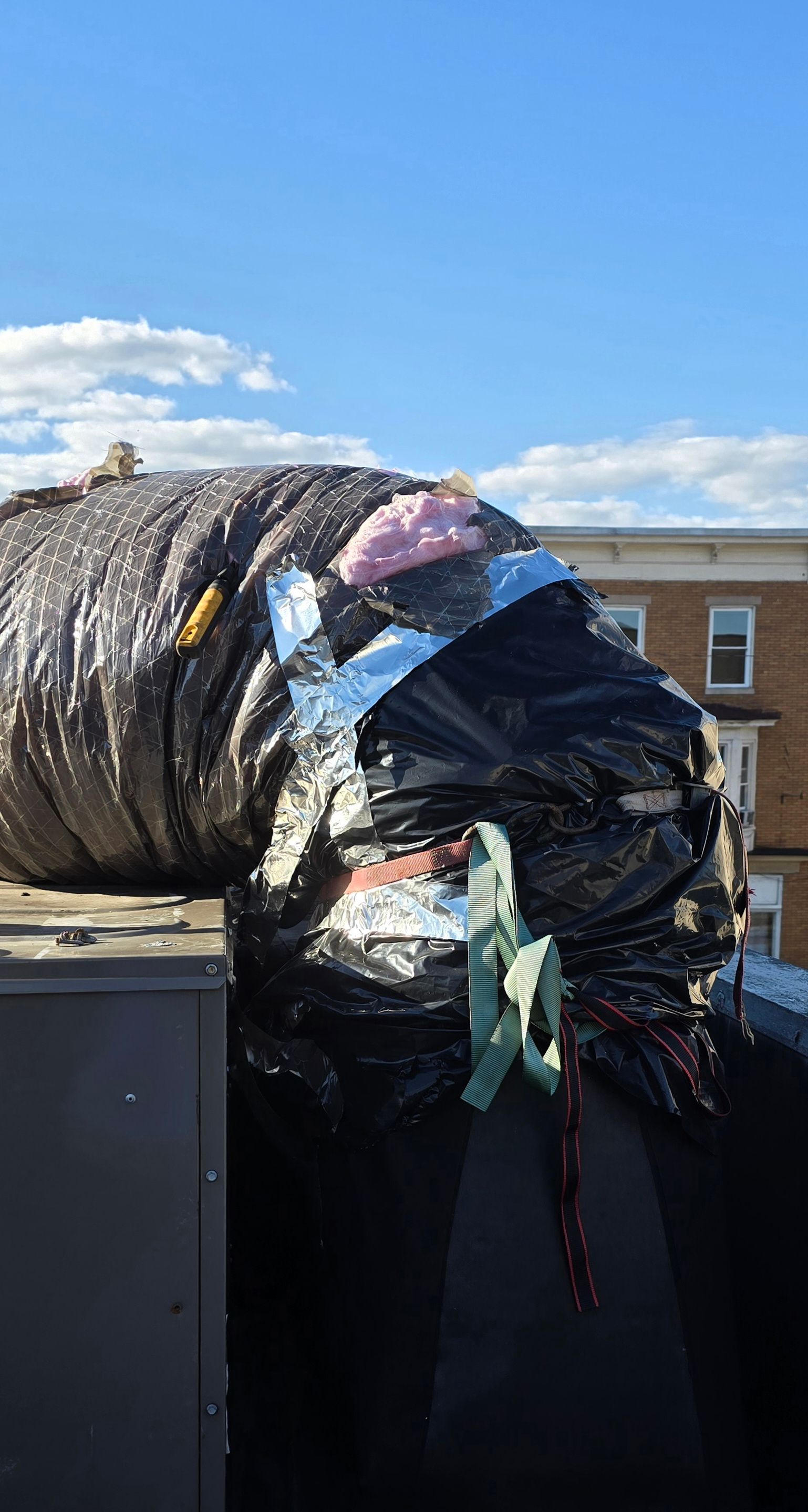 Large flexible air duct hose with silver and black insulation sitting on a rooftop under a clear blue sky.