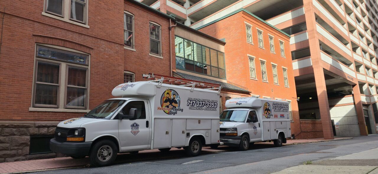 Two white work trucks are parked on a street in front of a brick building with a garage structure nearby.