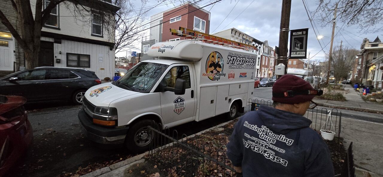 A person in a gray hoodie stands on a city street next to a white service van with a logo, parked by residential houses.