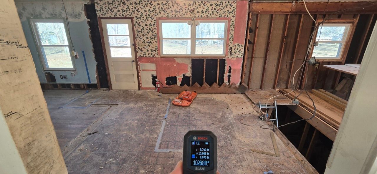 Hand holding a moisture meter in a home under renovation, showing exposed wall studs, subflooring, and a window.