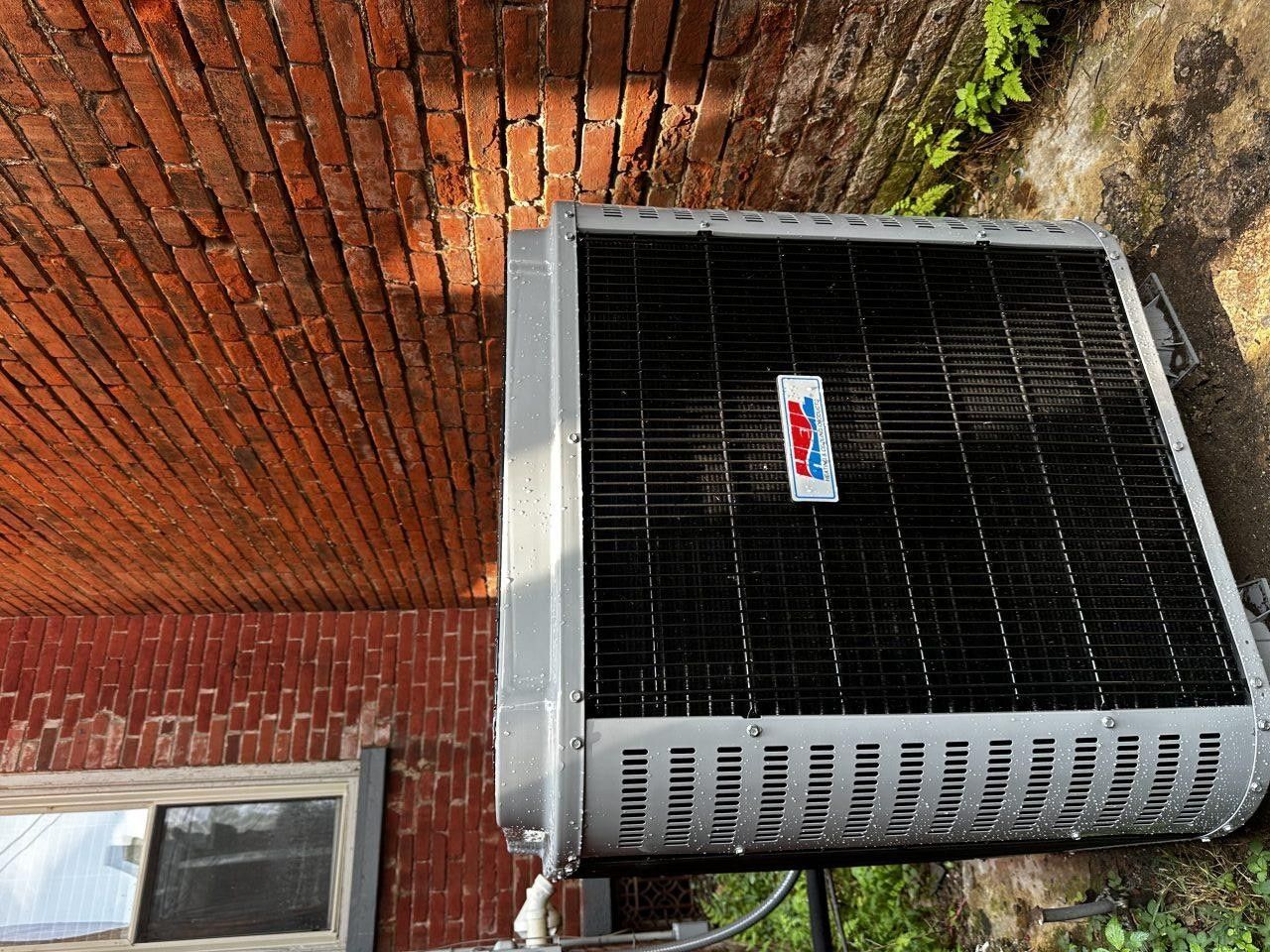 An outdoor air conditioning unit sits on the ground against a red brick wall.