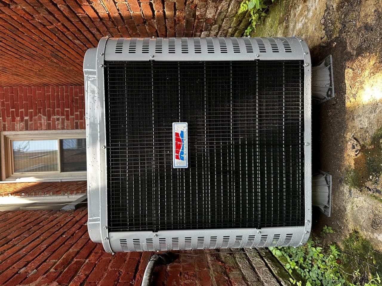 An outdoor air conditioning unit with a black grill and a white logo, positioned against a red brick wall.