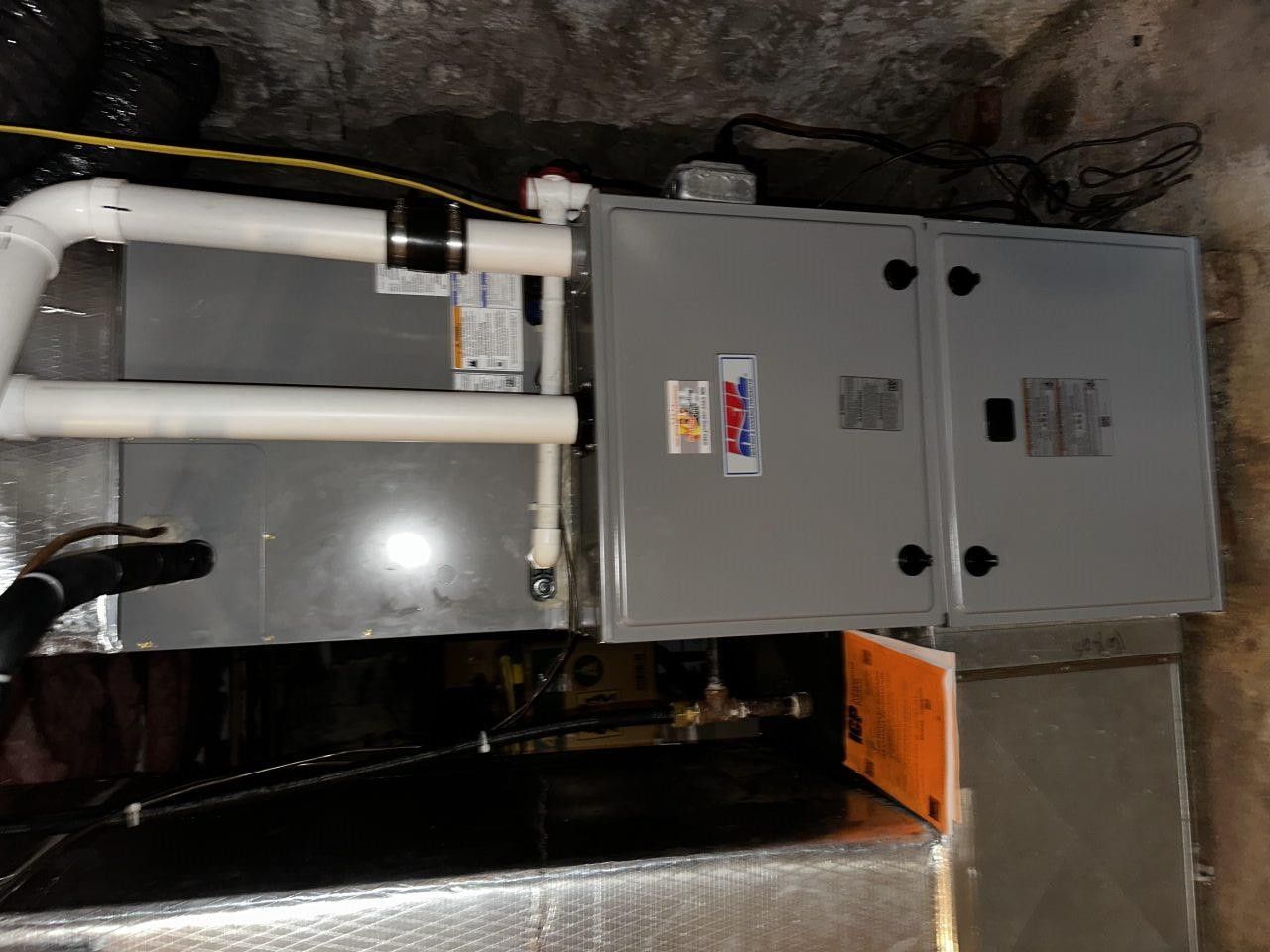 Gray HVAC furnace unit with white PVC intake and exhaust pipes installed in a basement.