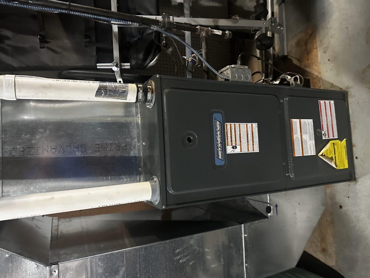 A gray residential furnace unit installed horizontally with white PVC piping attached to the side.