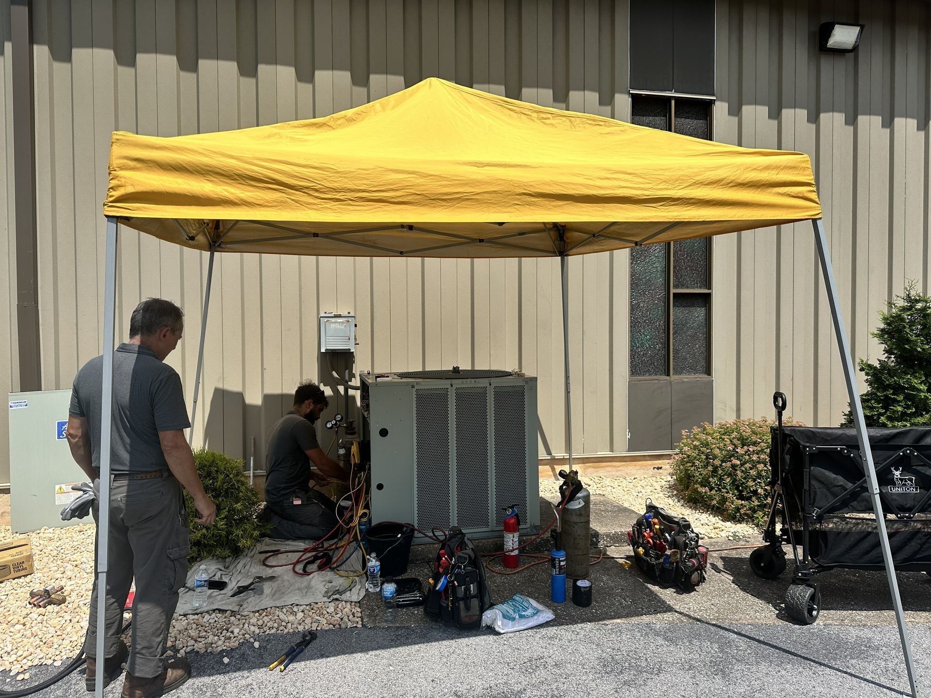 Two technicians work on an outdoor HVAC unit under a yellow canopy near a building wall.