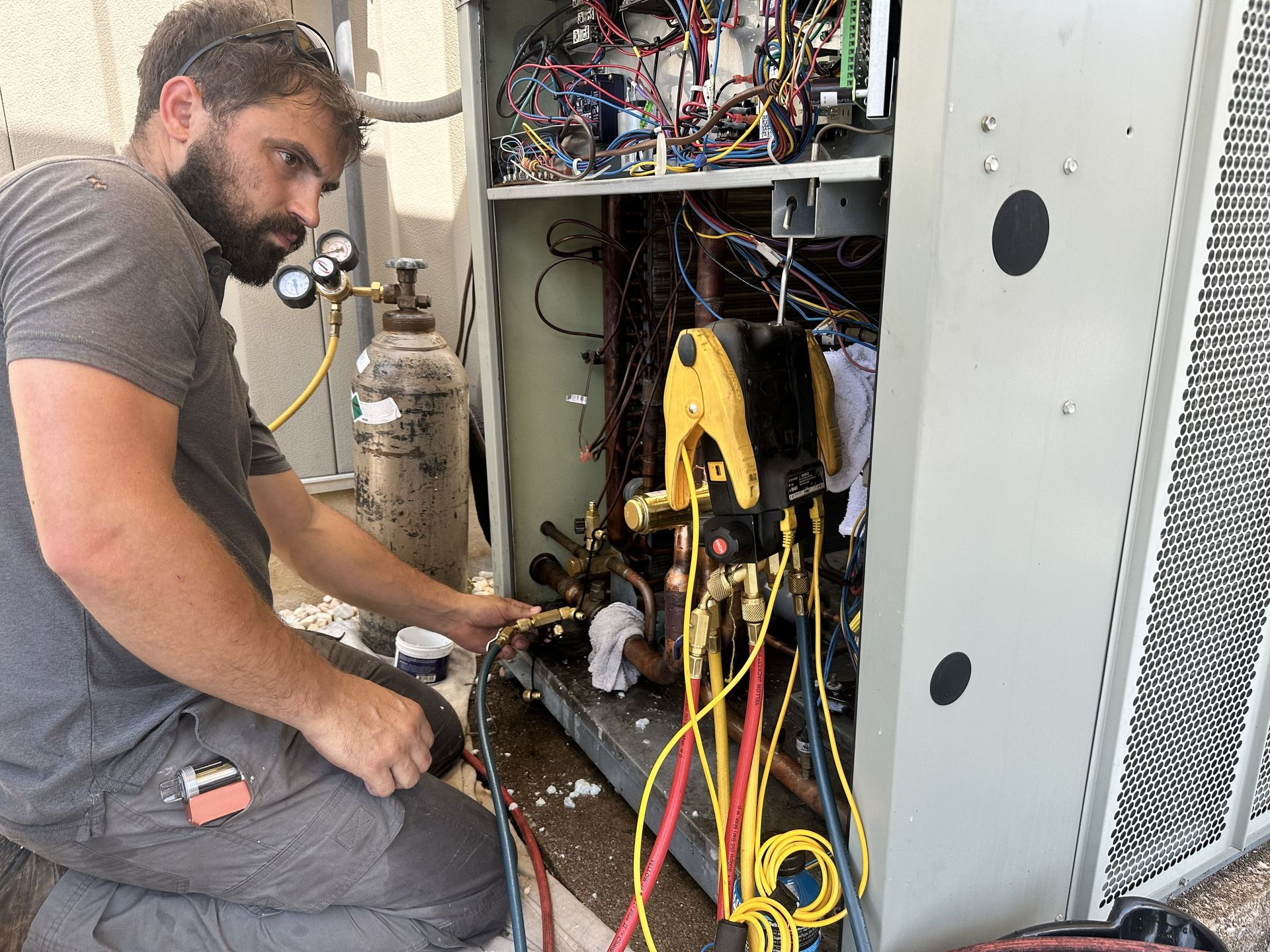 A technician kneels while repairing an HVAC unit, using gauges and a nitrogen tank to service the internal components.