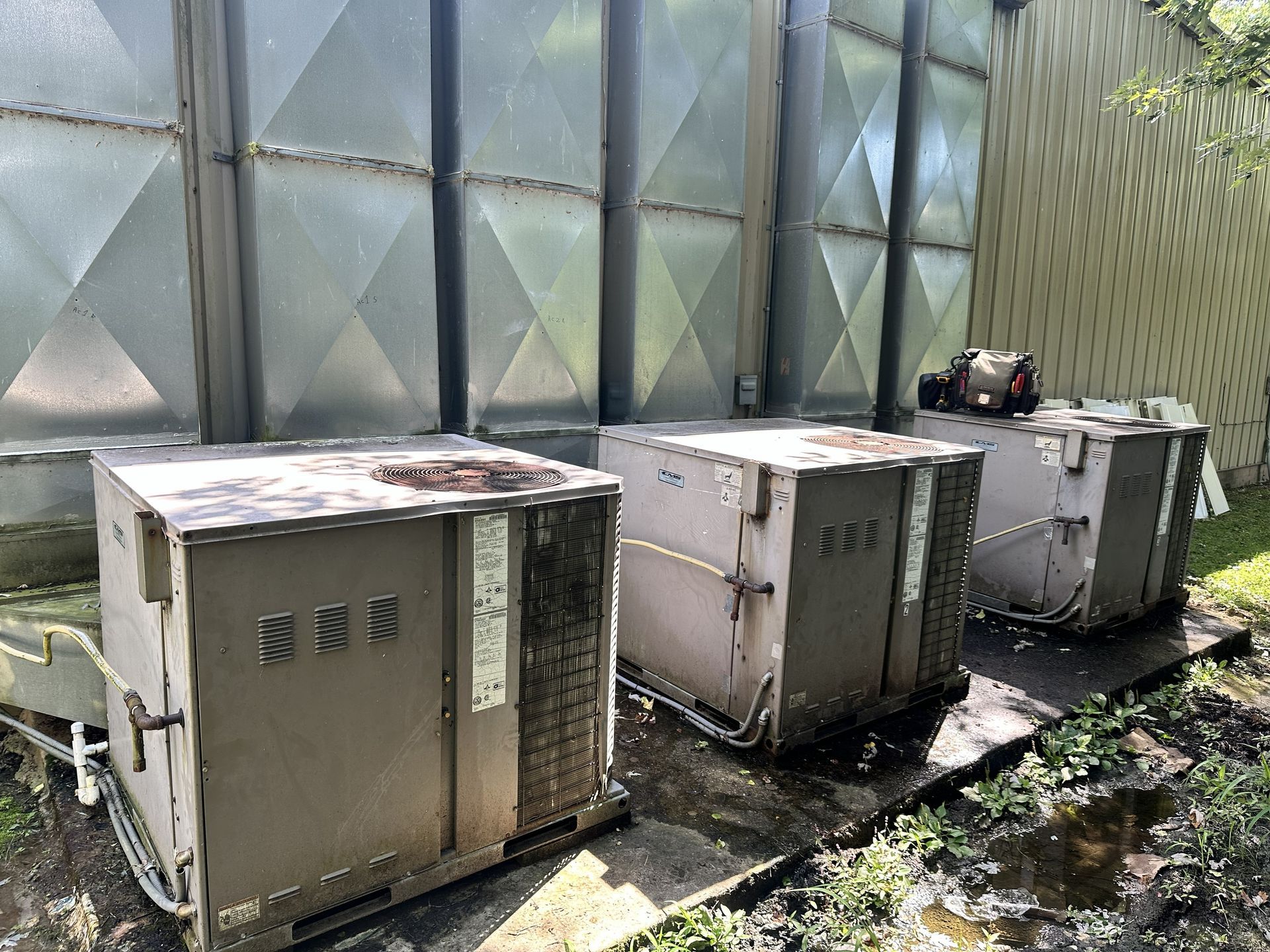 Three industrial air conditioning units positioned on a concrete slab in front of a metal-paneled wall.