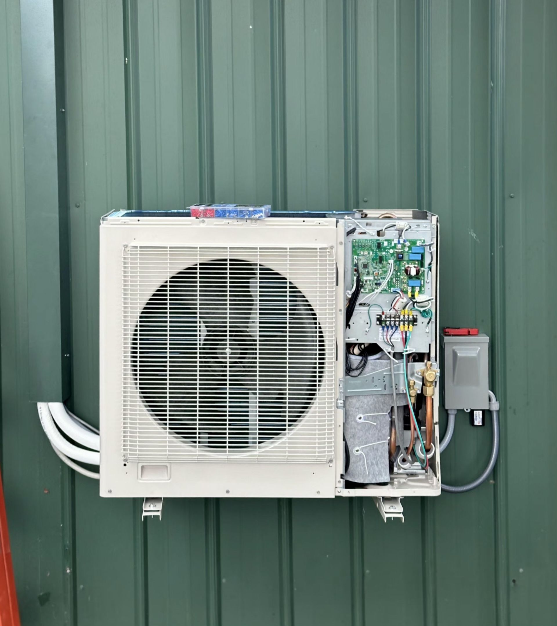 An outdoor white ductless mini-split HVAC unit mounted on a green metal wall with its electrical panel cover removed.