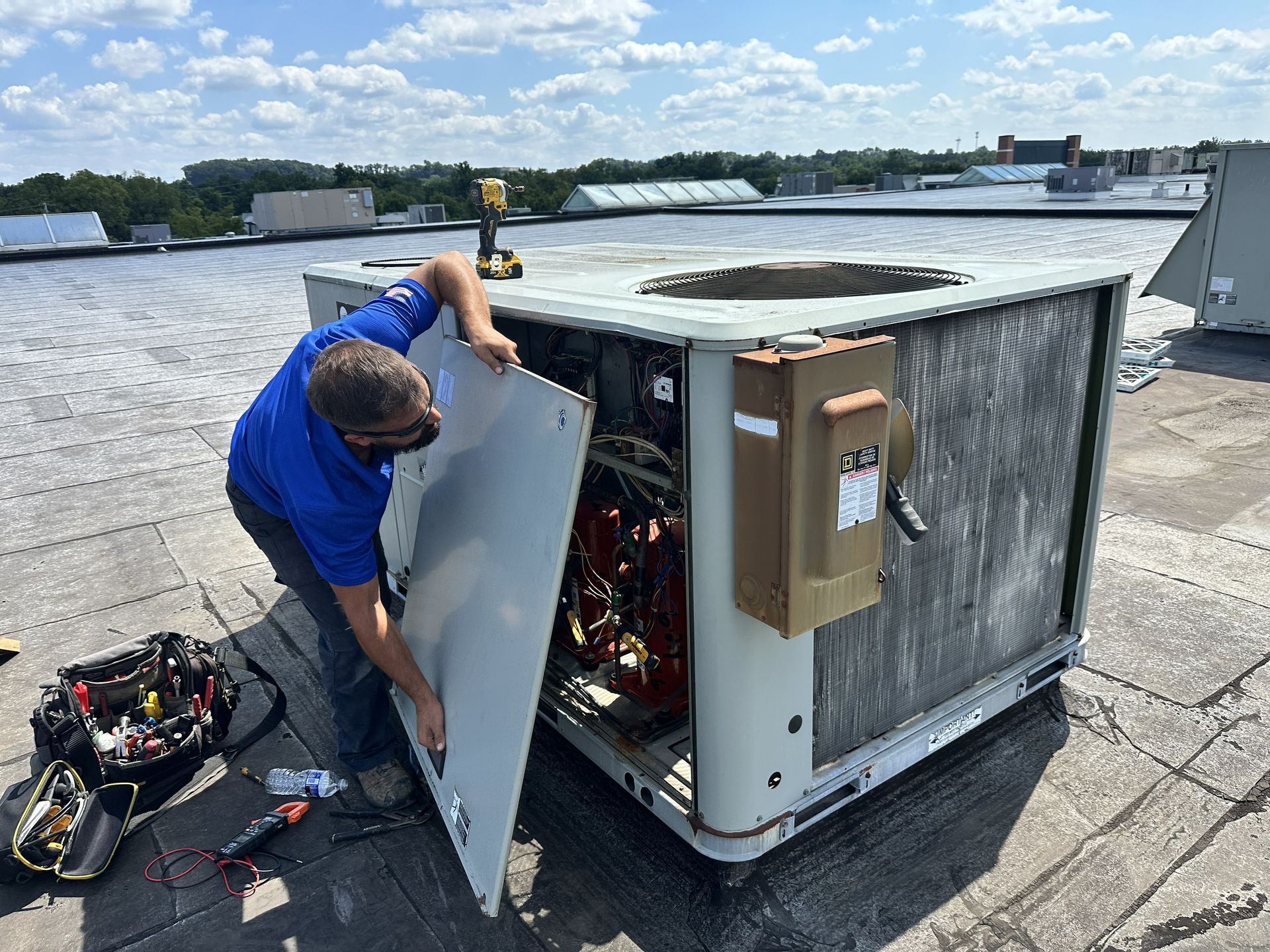 A technician in a blue shirt repairs a large HVAC unit on a flat rooftop on a sunny day.