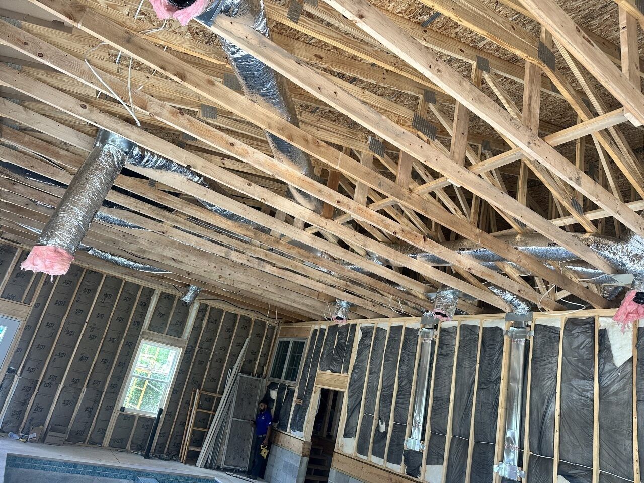An interior view of a room under construction with exposed wooden ceiling trusses, foil-covered air ducts, and insulation.