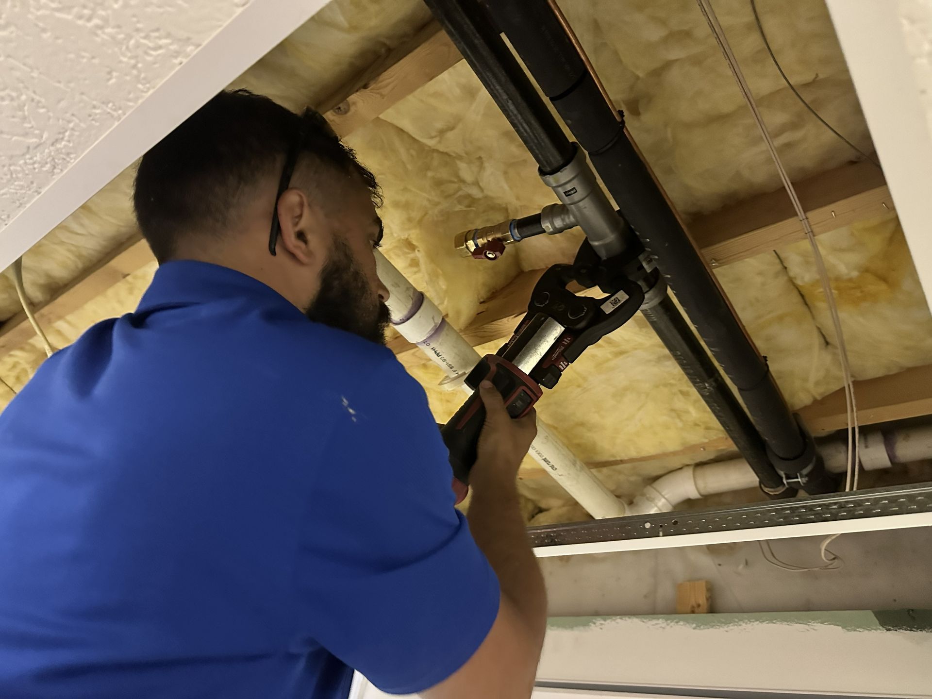 A person in a blue shirt uses a pipe-crimping tool on metal pipes in an open ceiling with exposed insulation.