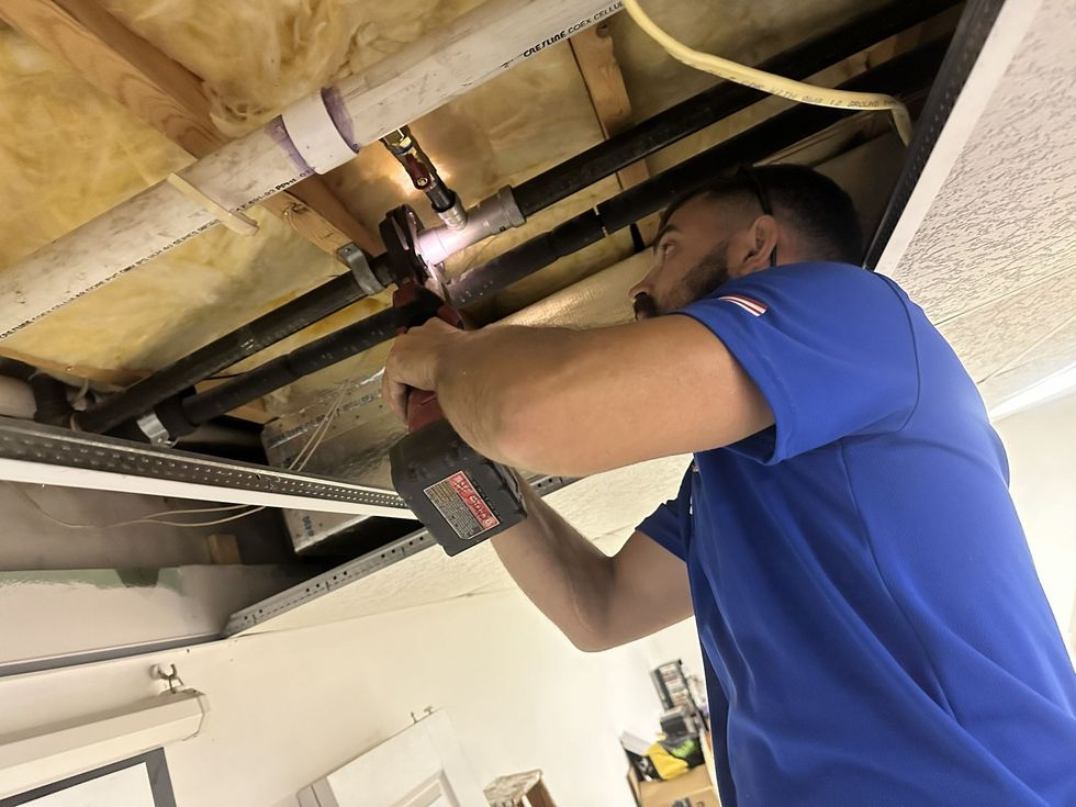 A worker in a blue shirt uses a power tool to install or repair plumbing pipes mounted to an open ceiling joist.