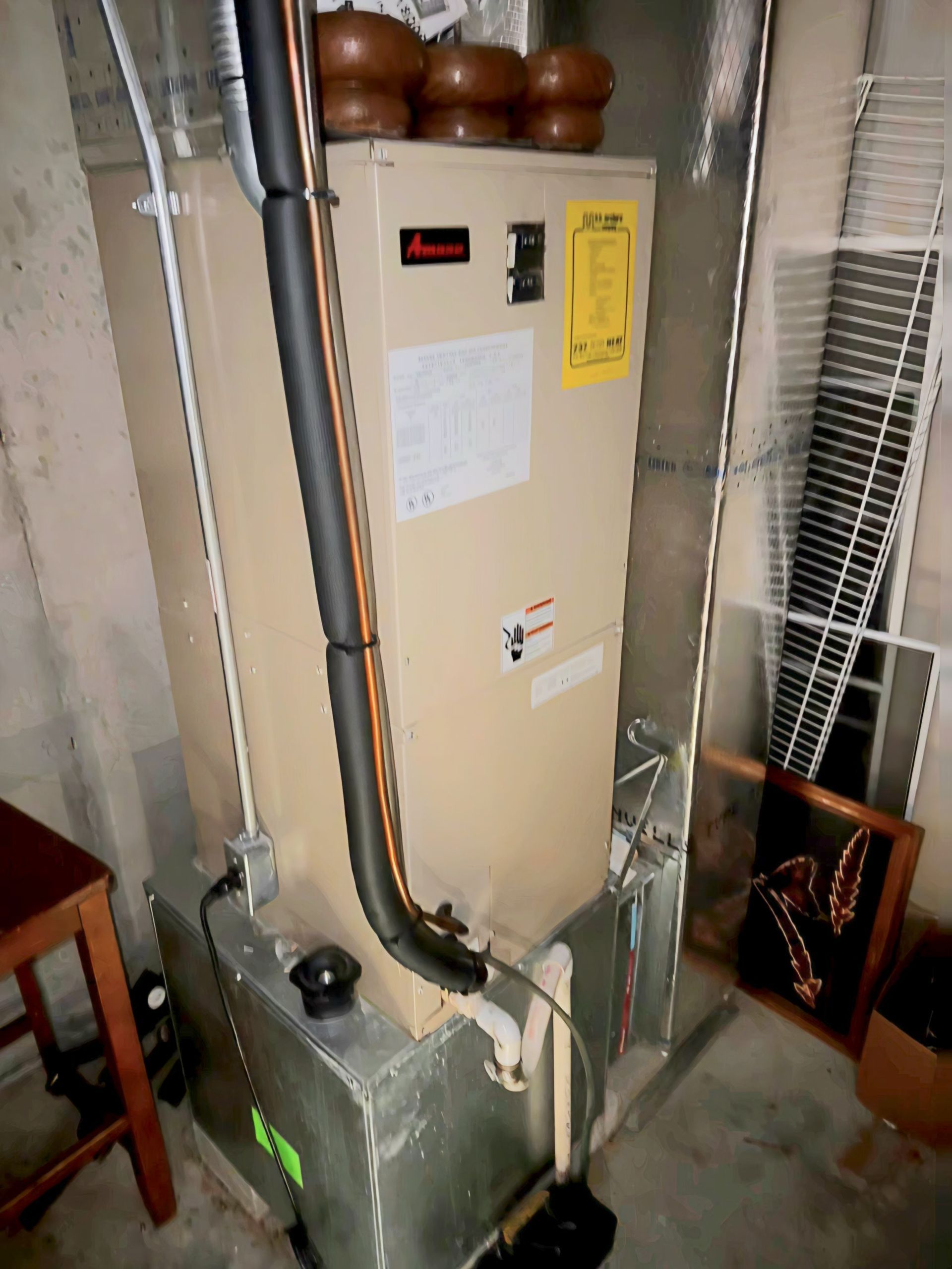 A beige Amana HVAC air handler unit standing upright in a basement corner next to metal ductwork.