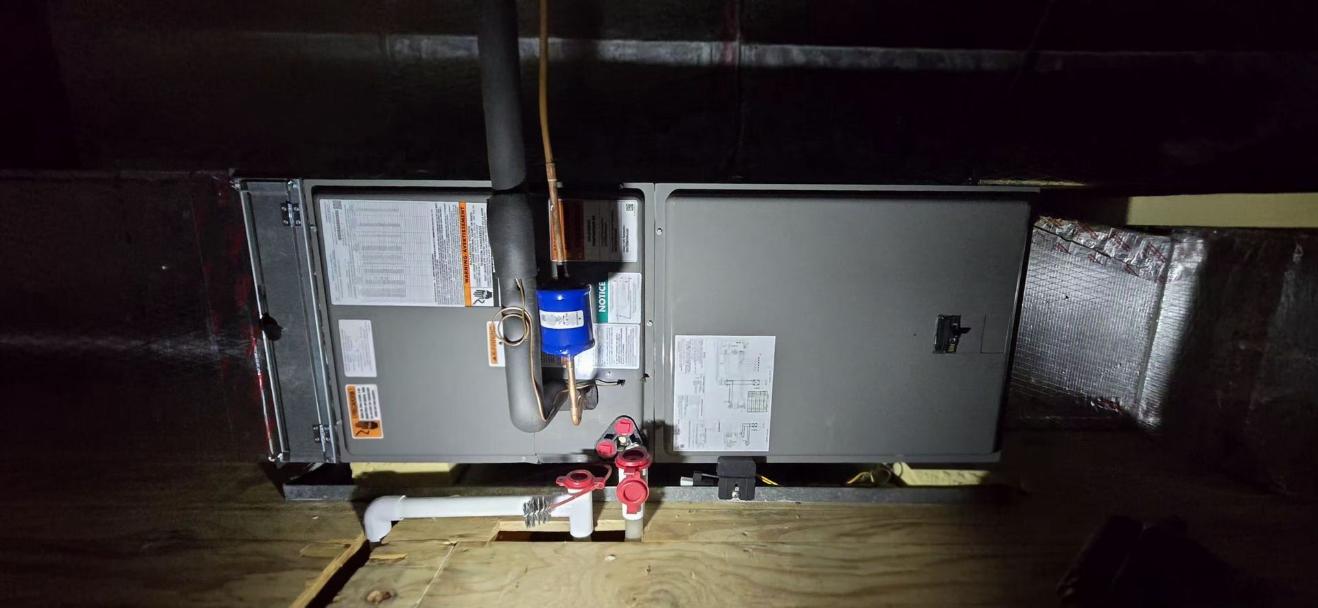 A grey HVAC air handler unit installed in an attic, with visible pipes, wiring, and a drain pan on the floor.