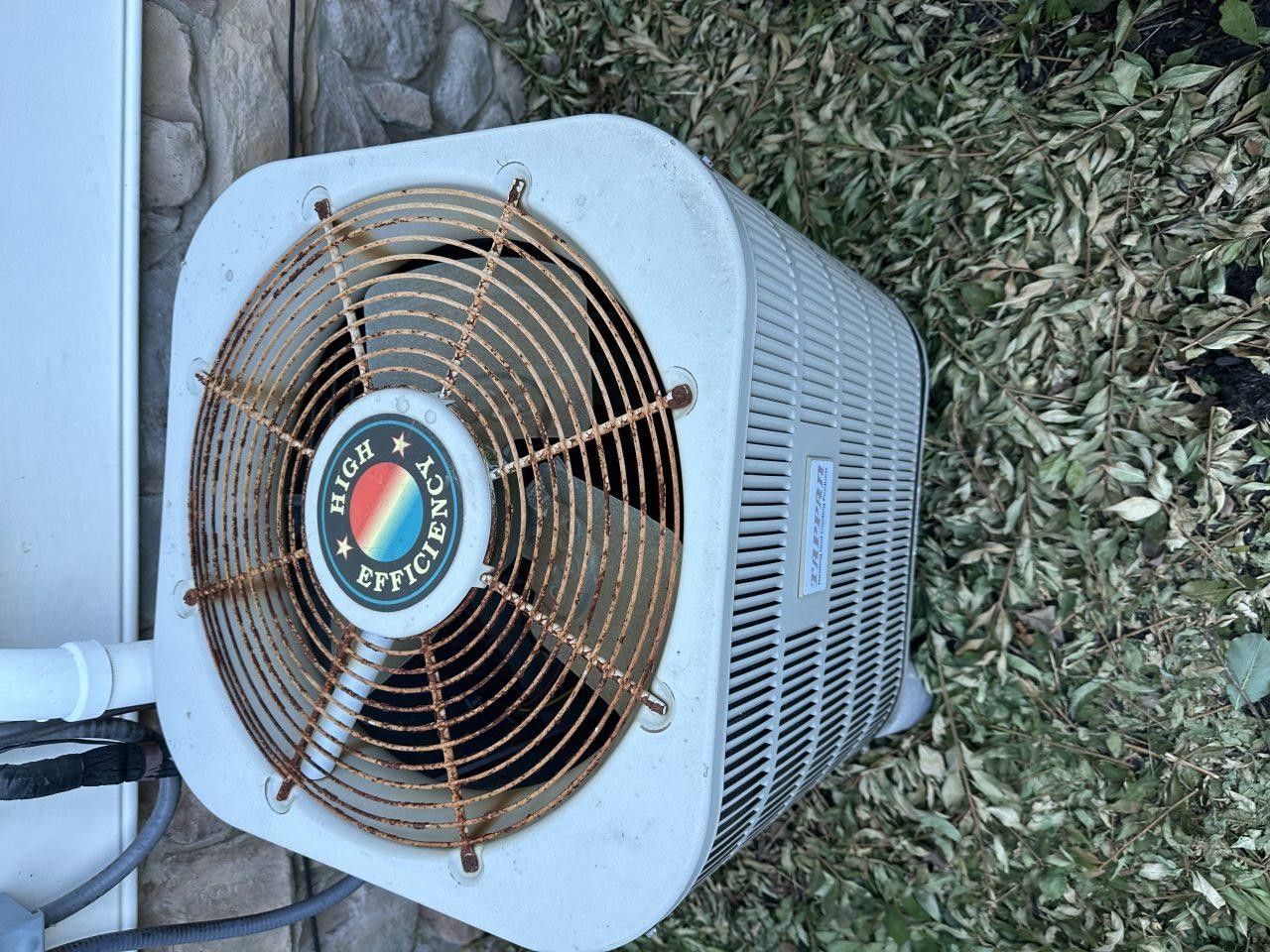 Top-down view of an outdoor air conditioning unit with a rusty fan grille, situated on the ground next to a wall.