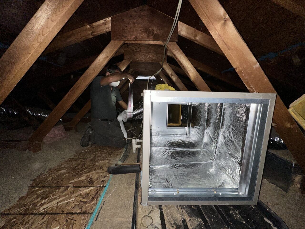 A technician in a mask works on HVAC equipment inside a residential attic with wooden roof trusses.