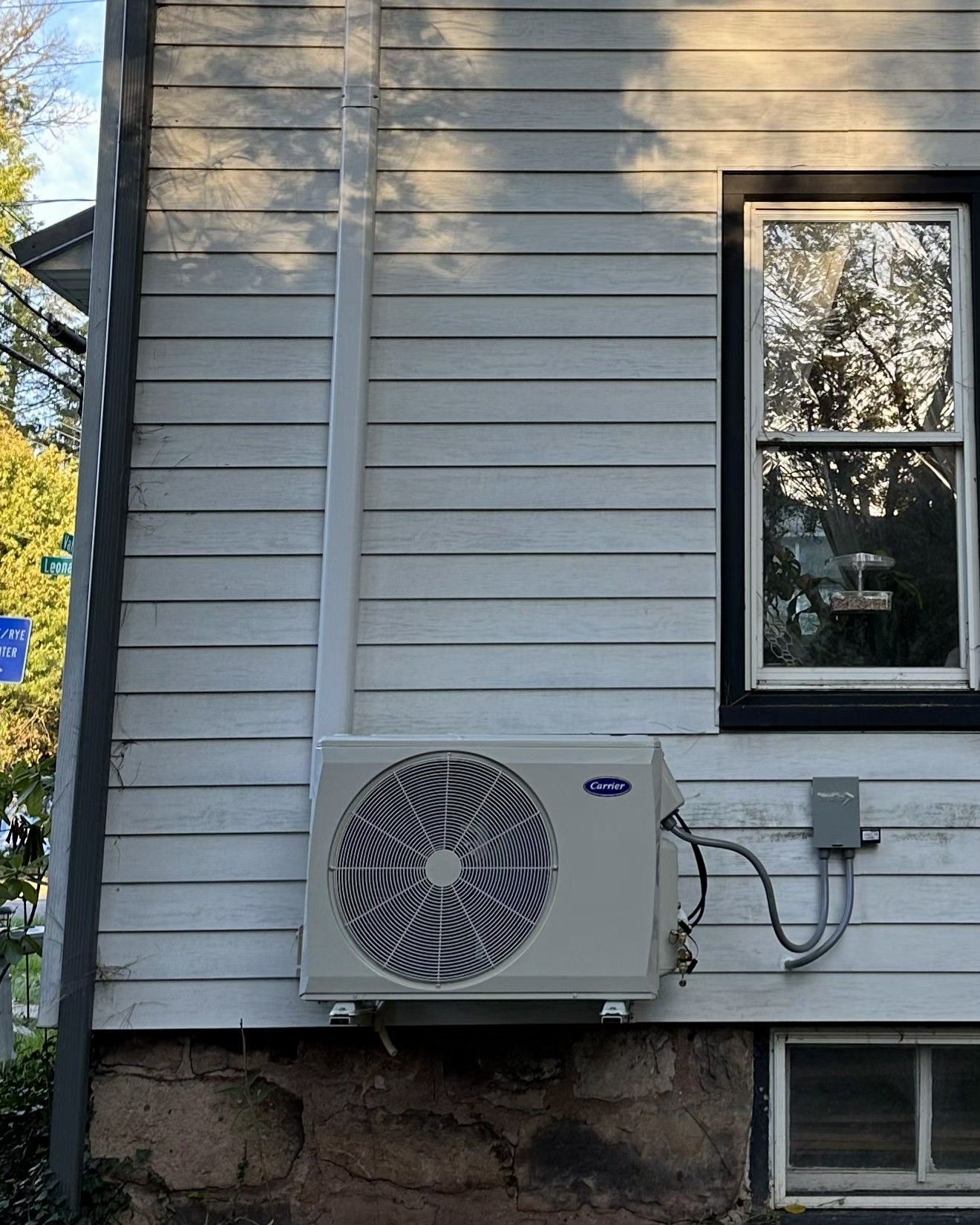 A white, wall-mounted outdoor mini-split AC unit on the exterior of a white-sided home near a stone foundation.