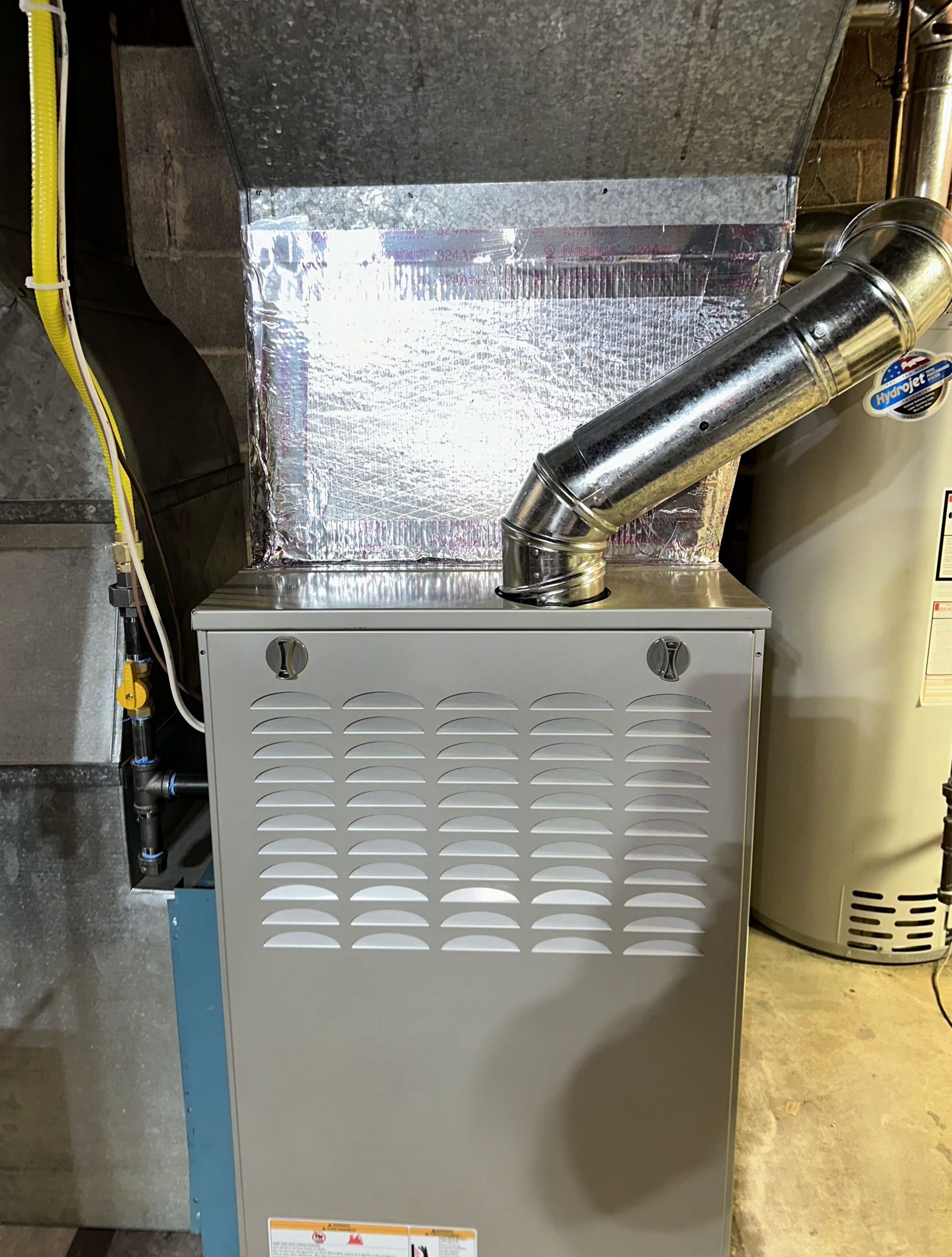 A beige residential gas furnace in a basement, featuring a metallic exhaust vent and foil insulation on the ductwork.