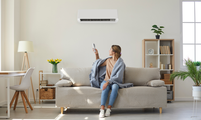 A person sitting on a sofa wrapped in a blanket, pointing a remote control at a wall-mounted air conditioner.