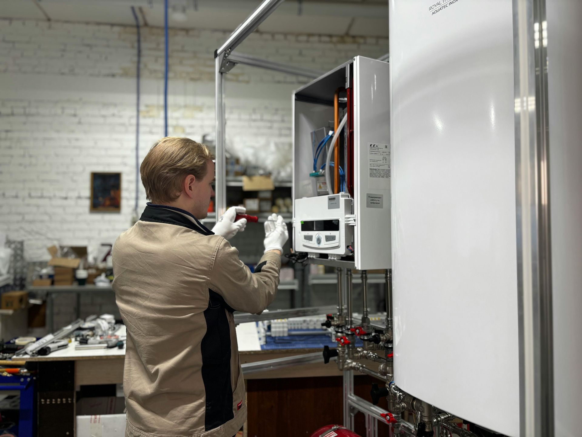A person in a beige shirt and white gloves works on the internal components of a wall-mounted heating unit in a workshop.