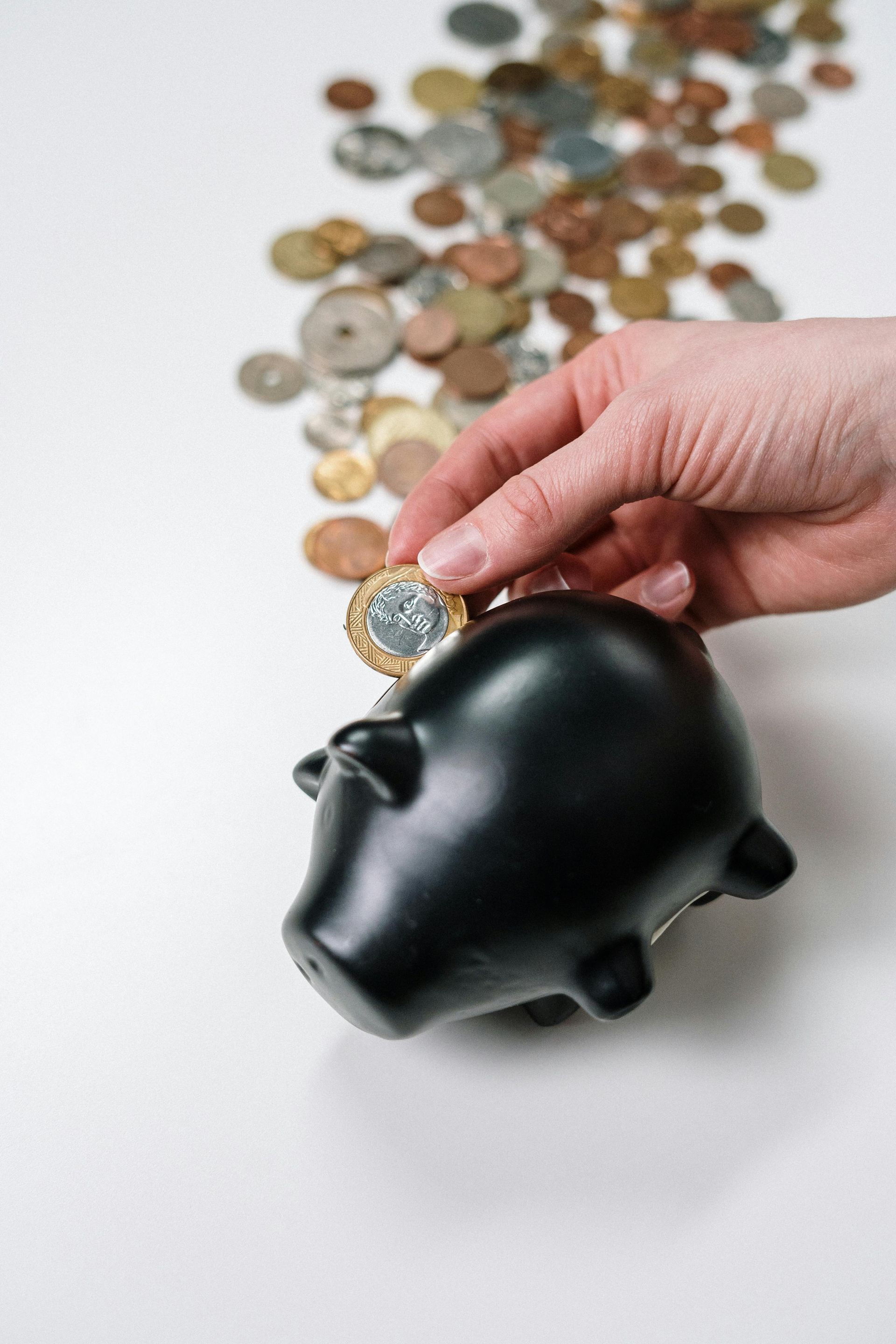 A hand places a metallic coin into a black piggy bank on a white surface scattered with many other coins.