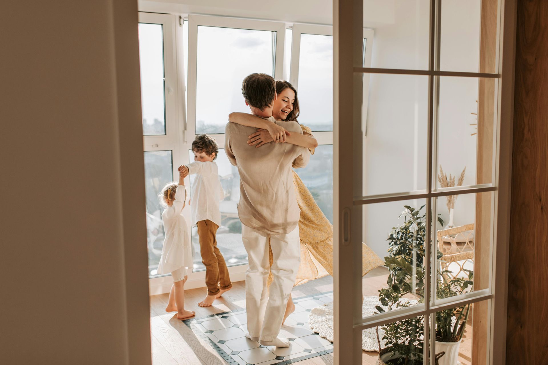 A family of four shares an affectionate embrace and dances together in a sunlit room by a large window.