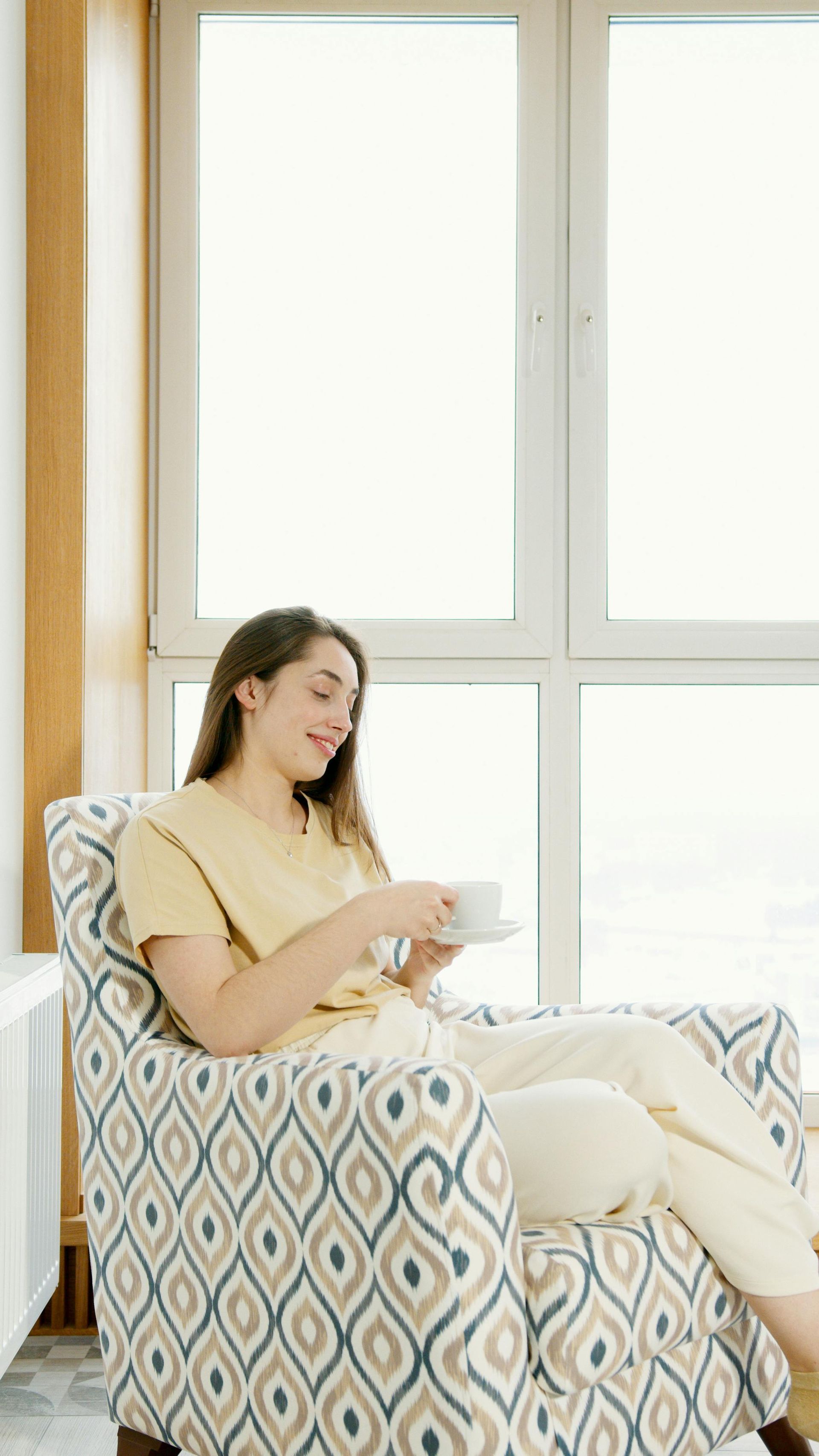 A person sits in a patterned armchair by a large window, holding a white mug and looking down with a gentle smile.