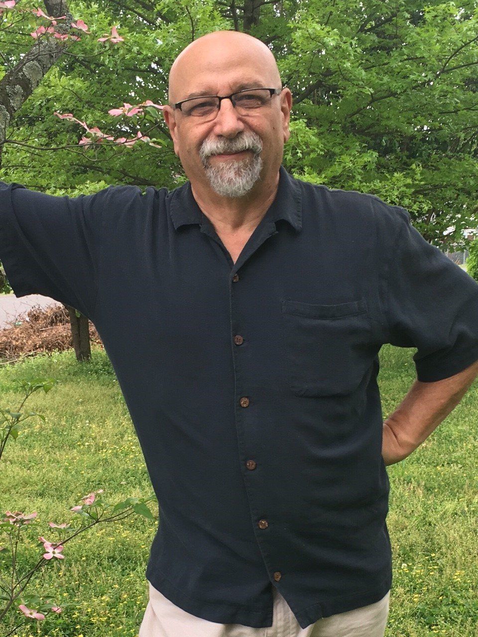 A man with a beard and glasses is standing in the grass with his hands on his hips.