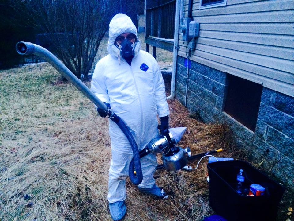 A man in a protective suit is holding a vacuum cleaner in front of a house.