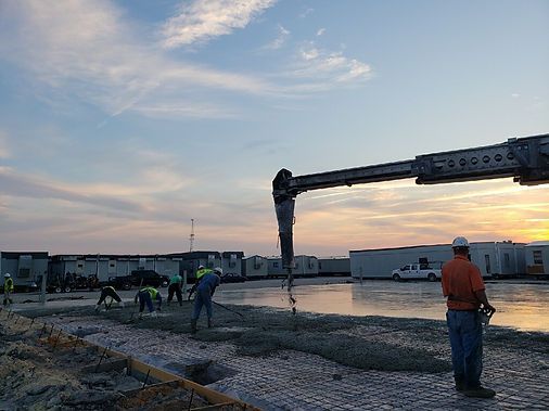 Construction workers pour concrete from a crane nozzle onto a mesh-covered foundation at sunset.