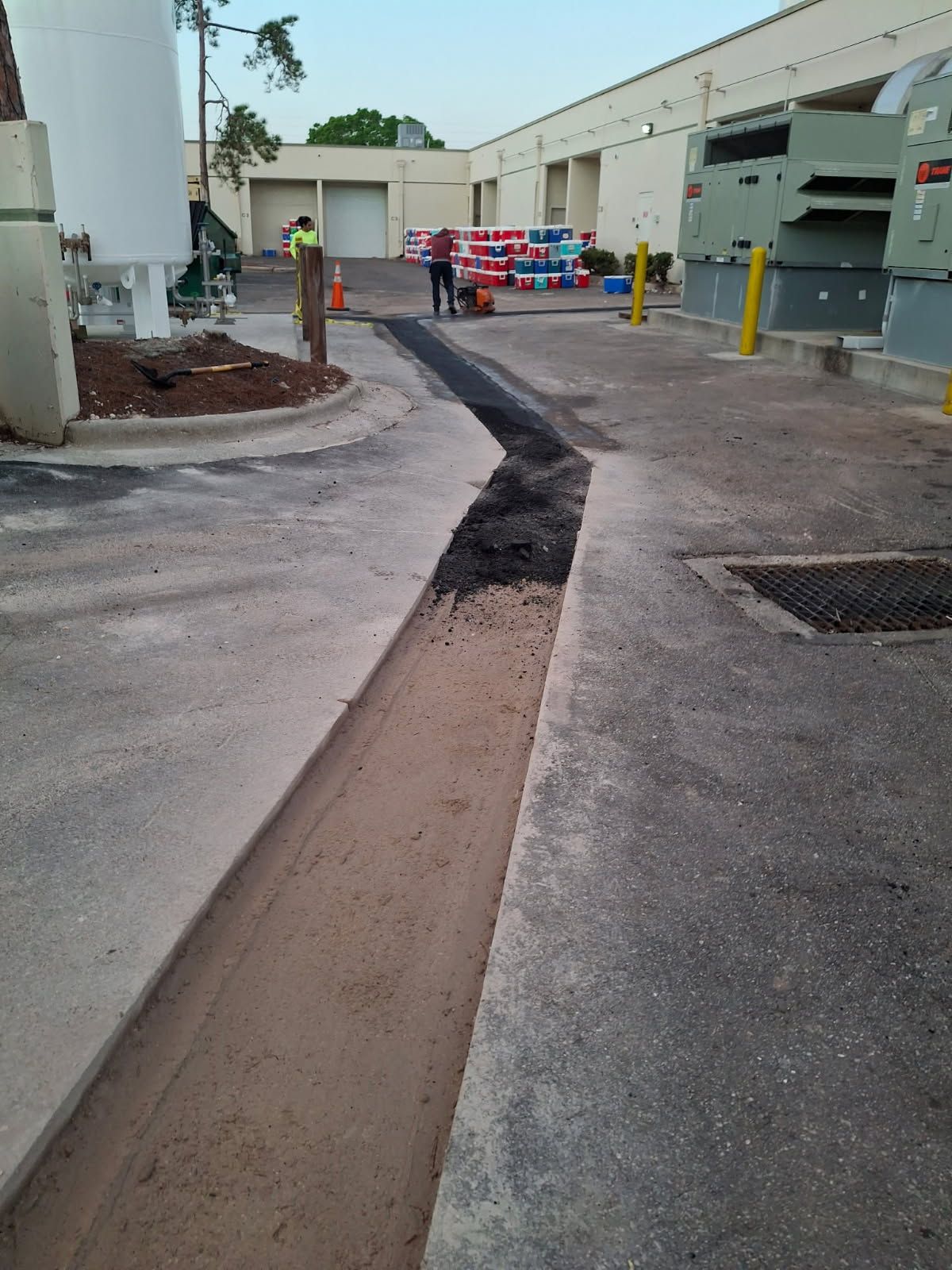 A construction worker uses a piece of equipment to fill a long, narrow trench in a paved industrial parking lot.
