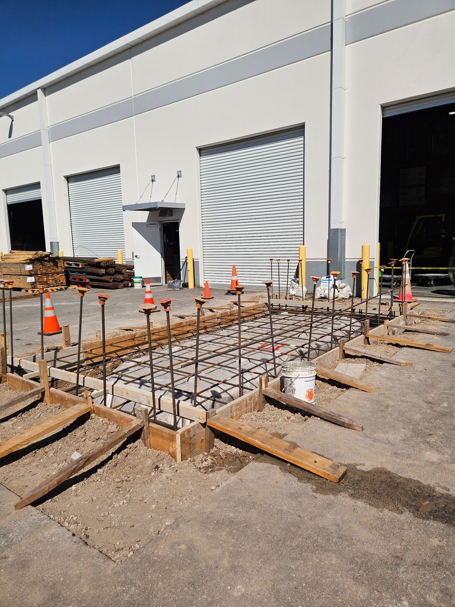 A rectangular concrete foundation form with metal rebar grid reinforcement sits in front of a white warehouse building.