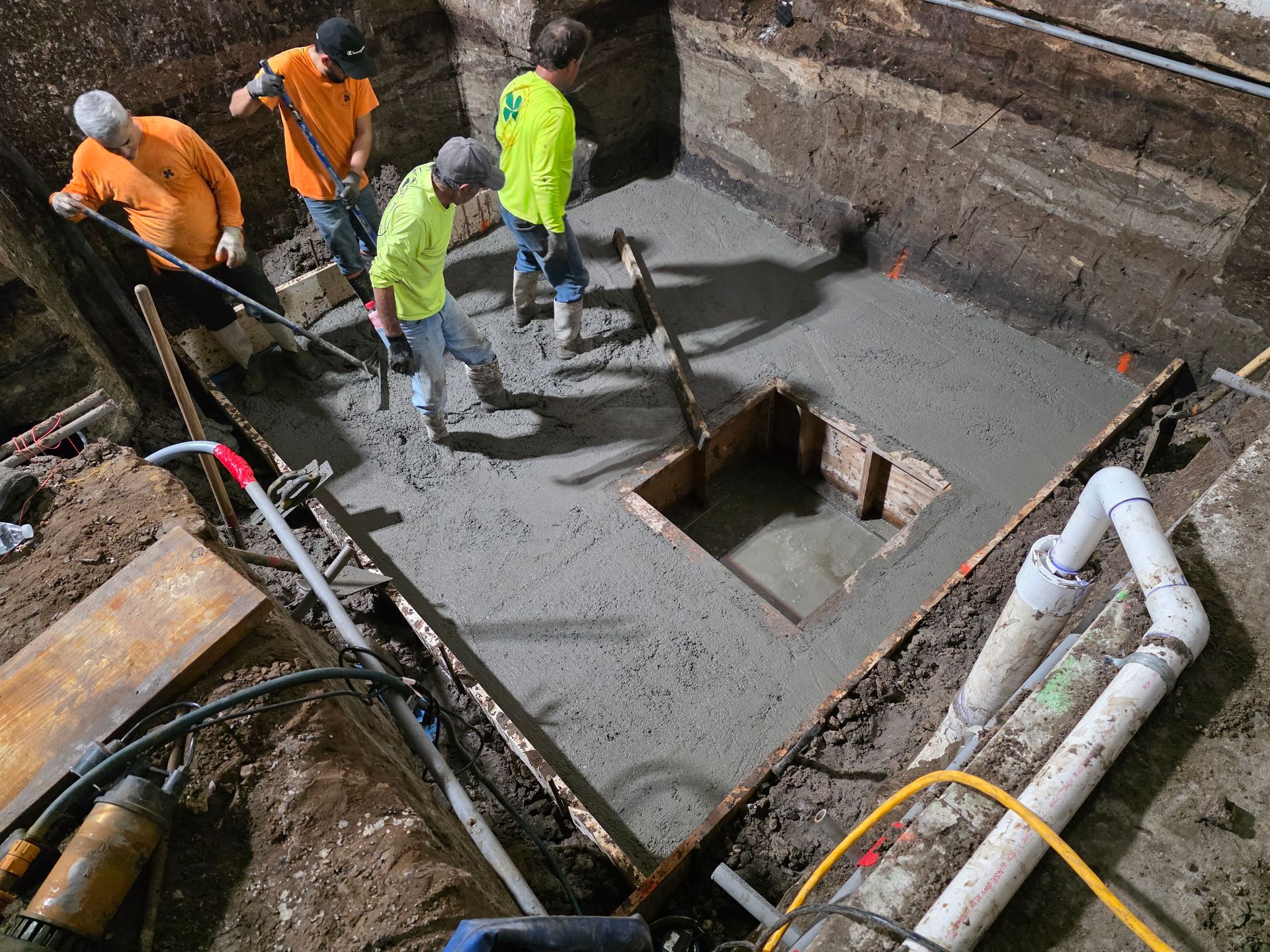 Four construction workers in work clothes smooth fresh concrete around a rectangular cutout in an excavated pit.