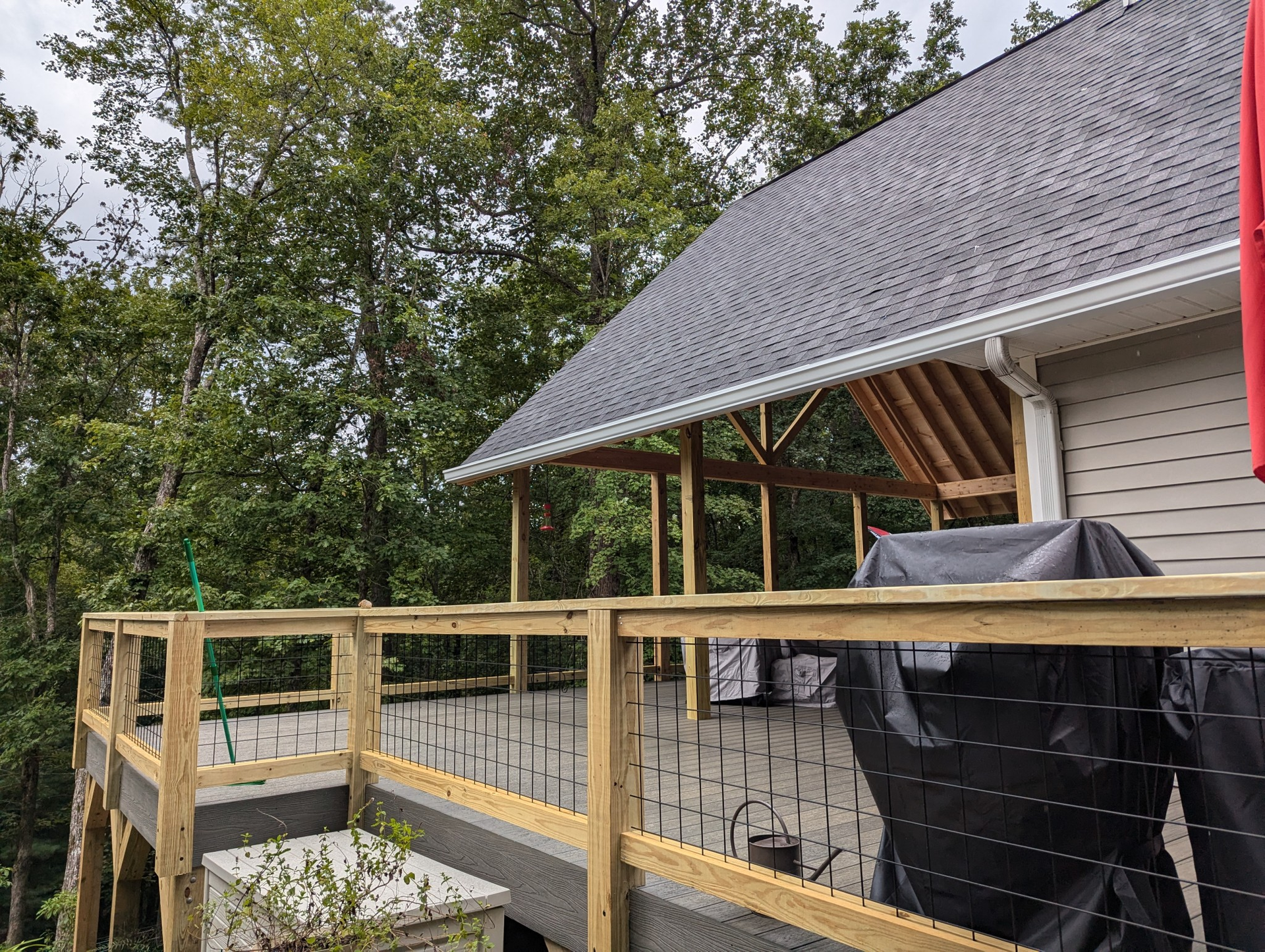 Wooden deck with railing attached to a house with a gray roof and trees in the background.
