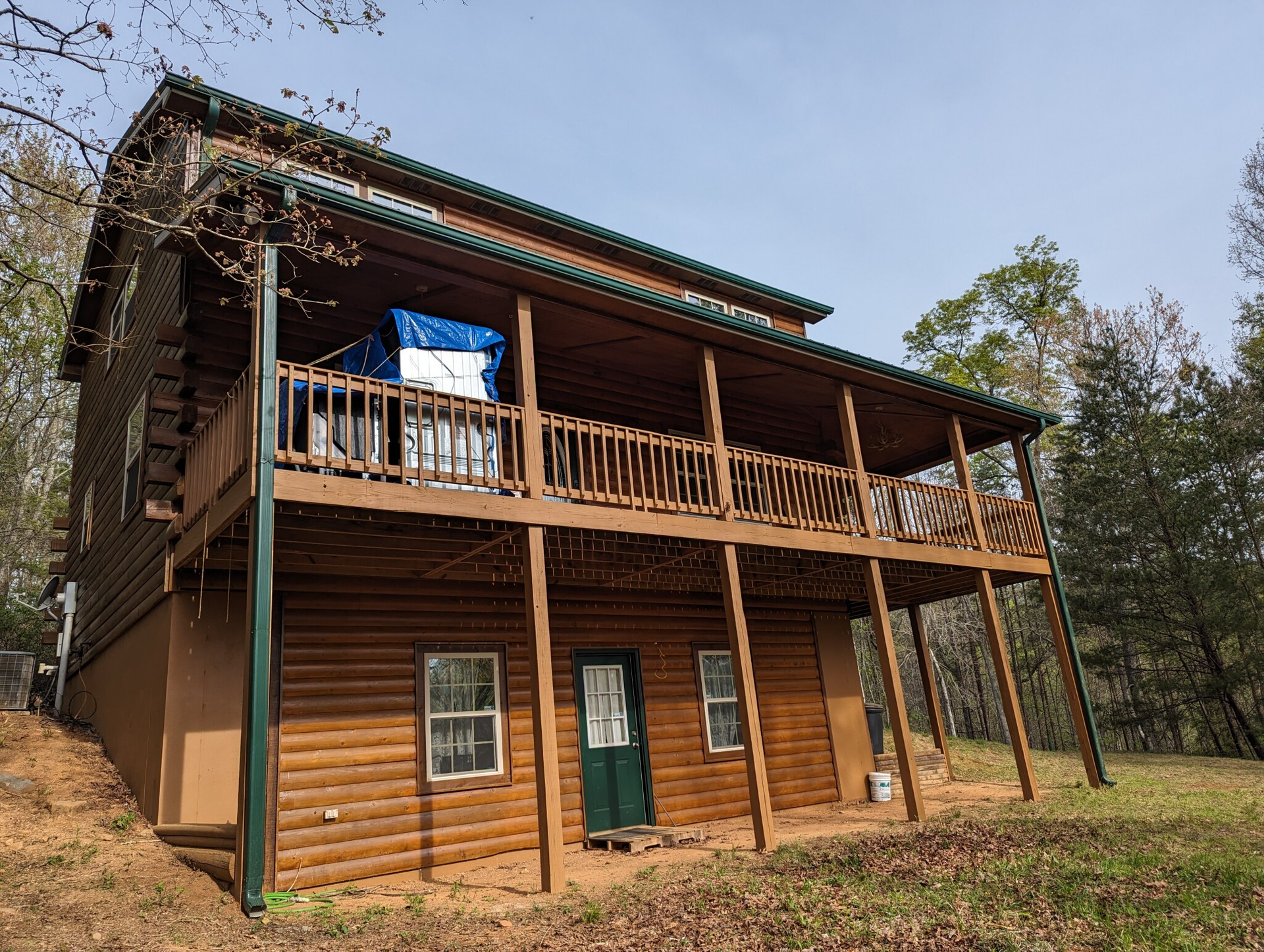 Two-story log cabin with a covered porch. Brown logs, green trim, blue tarp covers porch contents.