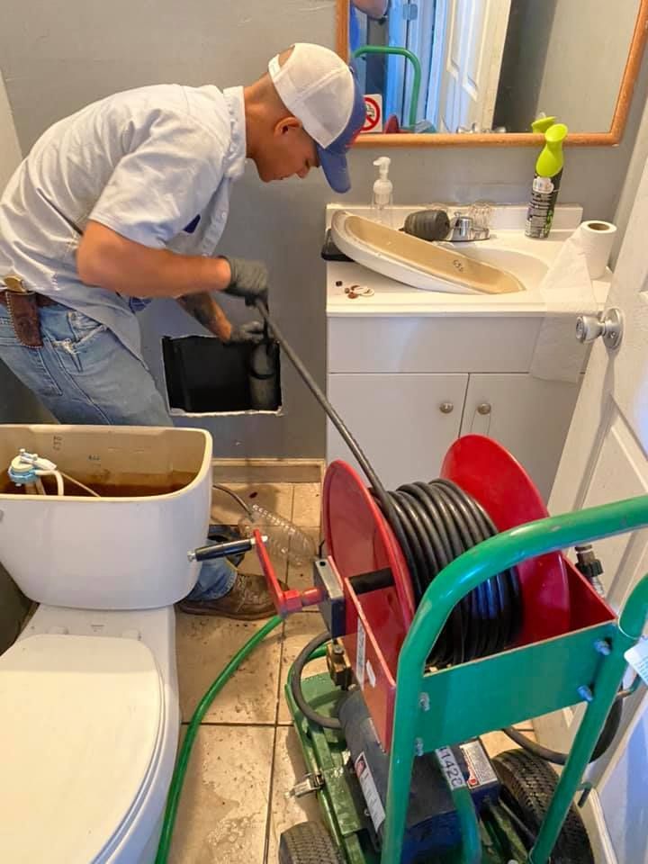 Plumber using a drain snake under a bathroom sink, with tools and hose reels on the floor.