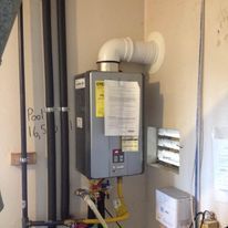 Wall-mounted gray tankless water heater with white vent pipes and nearby electrical boxes in a utility room