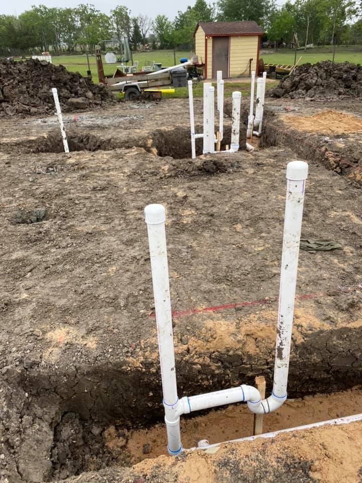 PVC pipe stubs in a dirt foundation trench at a construction site, with a small shed in the background