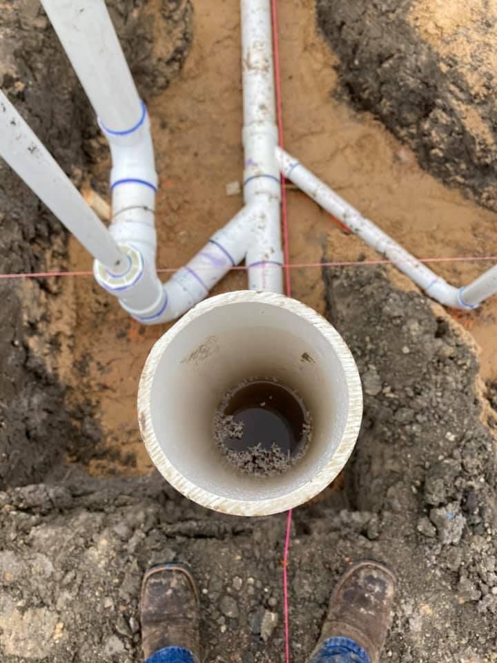White PVC pipes connected to a round pipe opening in a dirt trench.