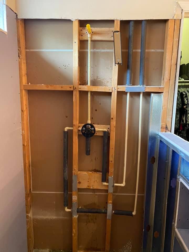 Wall framing with exposed plumbing and valves in an unfinished bathroom, with a blue bathtub on the right.