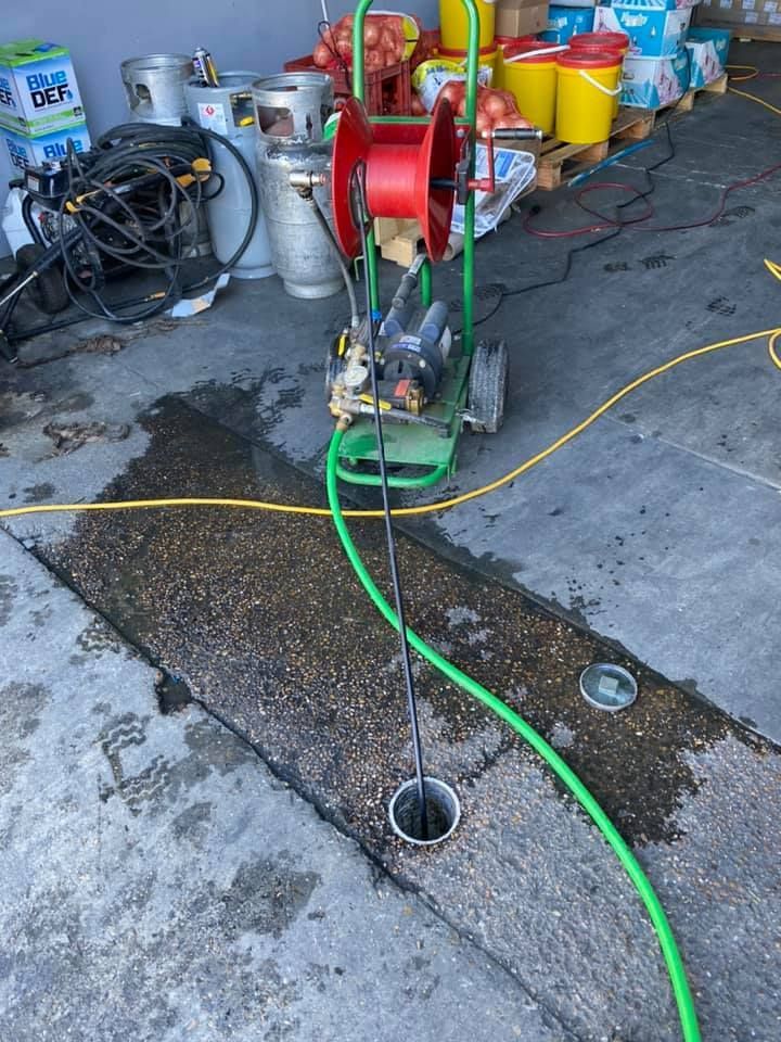 Green hose on a wheeled drain-cleaning machine in a garage, draining into a floor drain with tools nearby