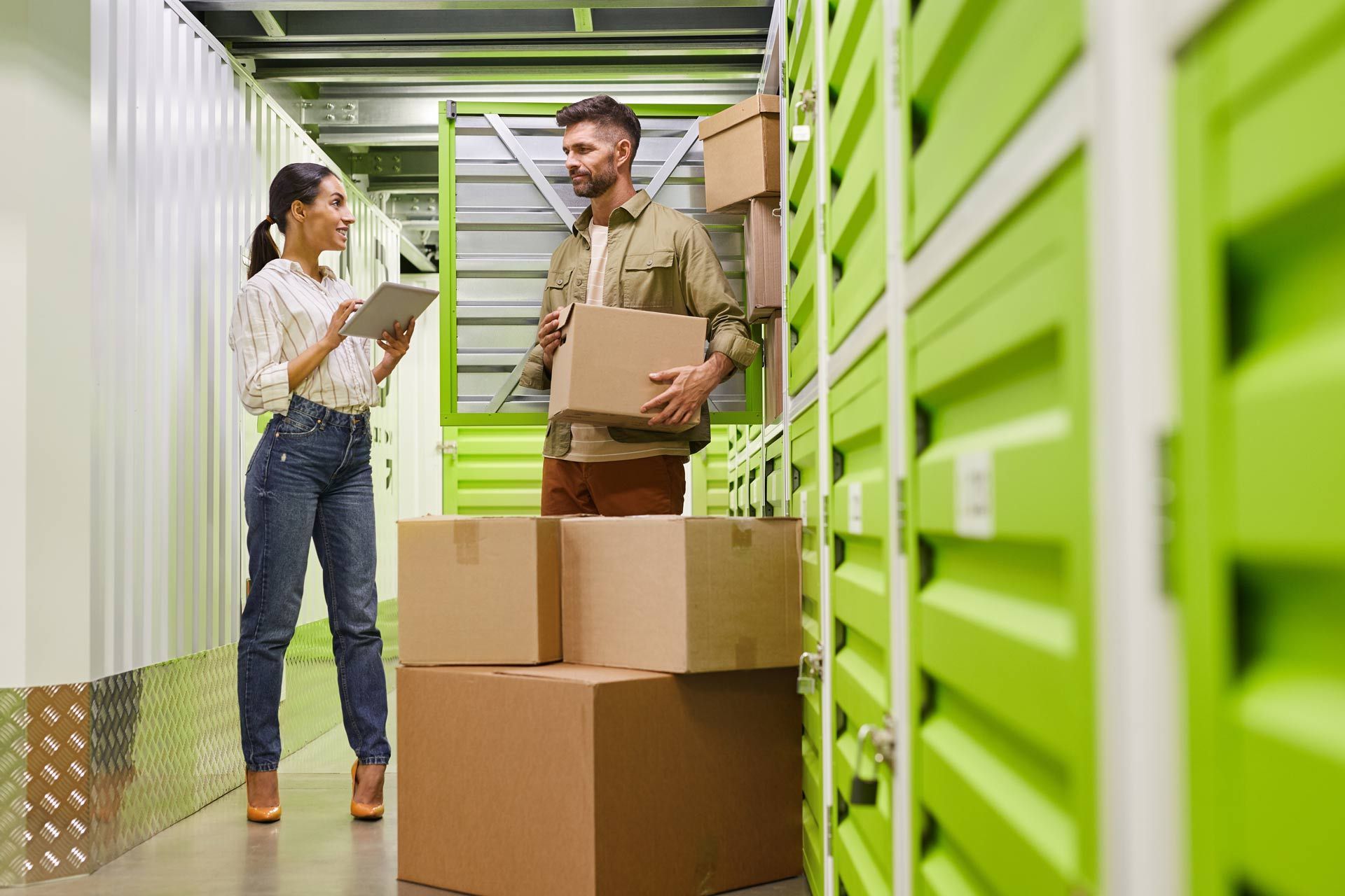 Woman with tablet and man holding a box in a storage unit; green lockers in the background.