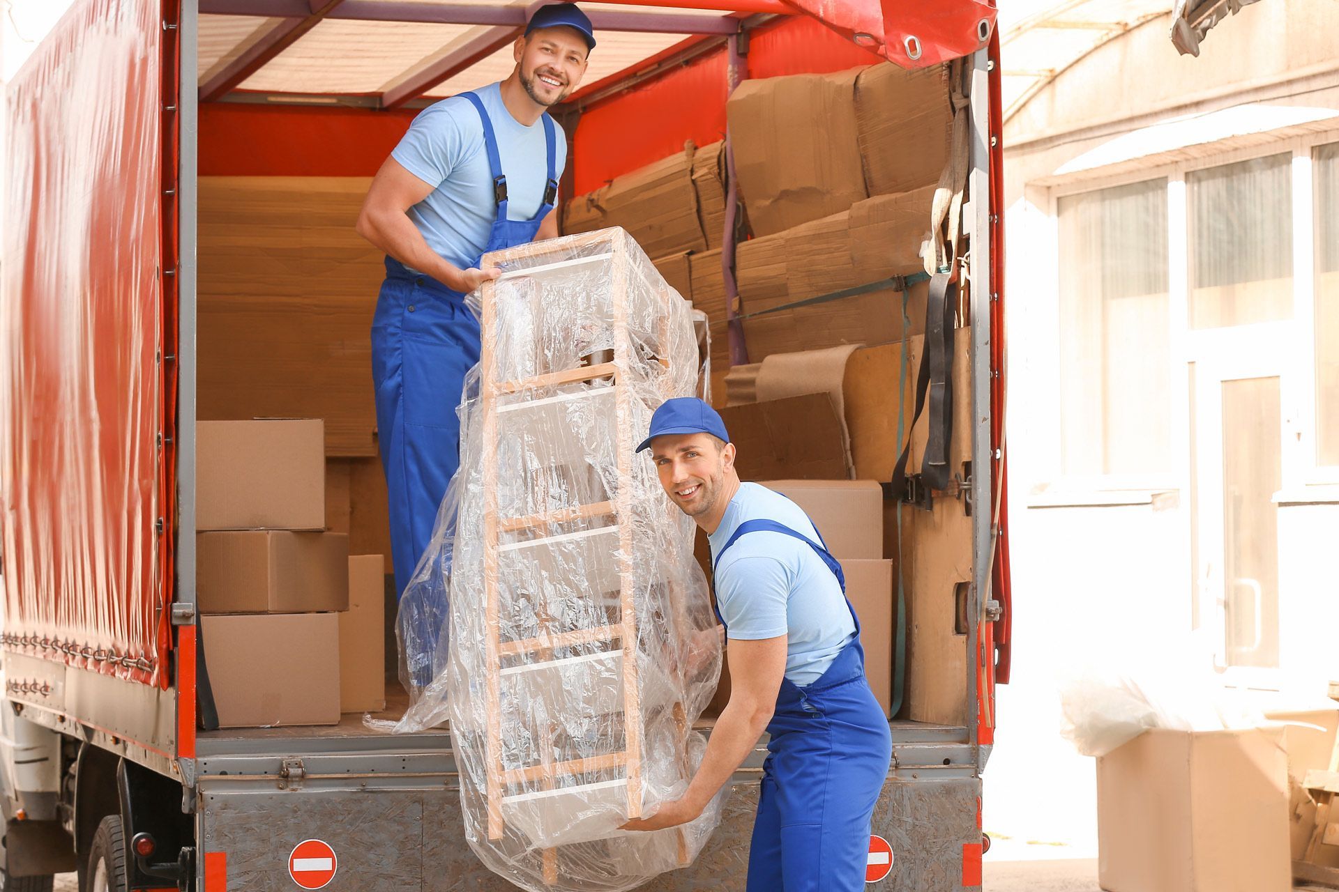 Two movers in blue overalls load a wrapped shelf into a red truck; they both smile.