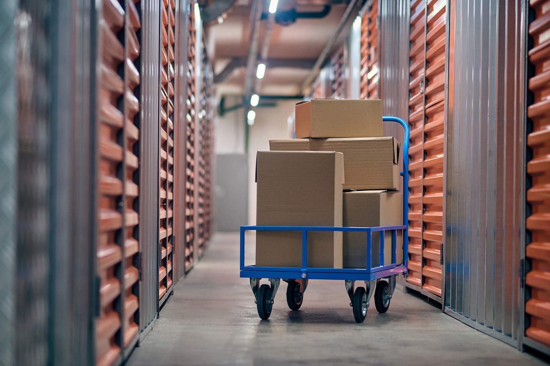 Blue cart loaded with cardboard boxes in storage unit hallway, orange units.