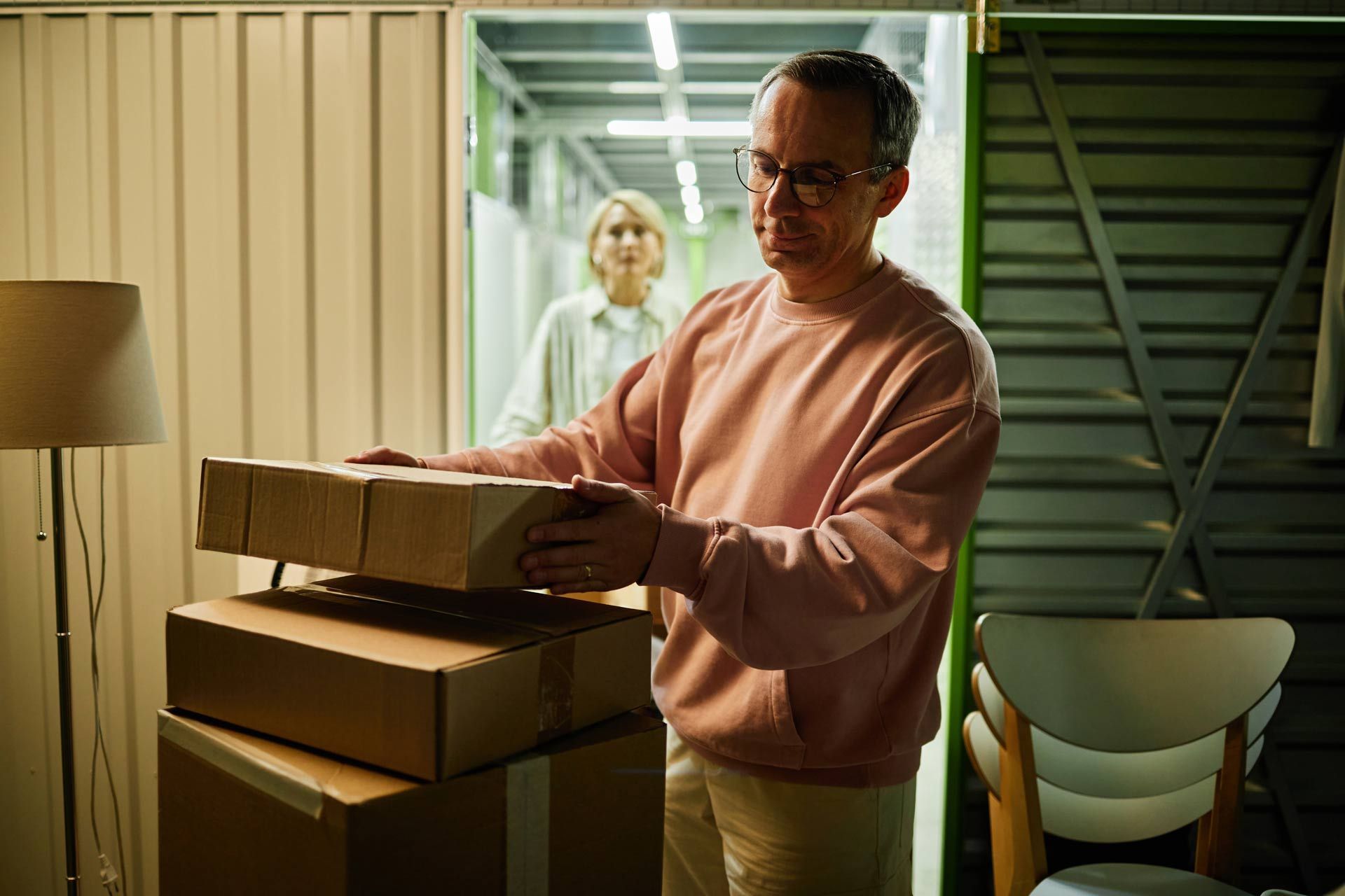 Man carrying stacked boxes in a storage unit, woman in background.
