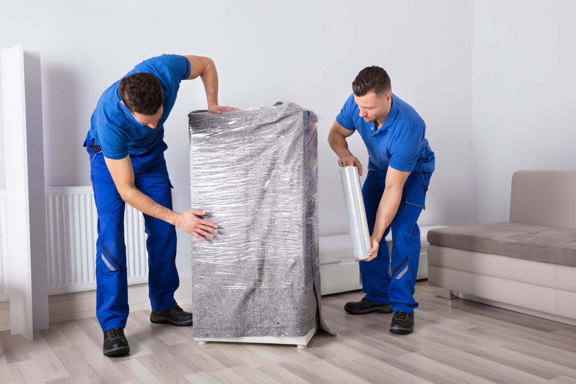Two movers in blue uniforms wrap a white cabinet with plastic wrap in a living room.
