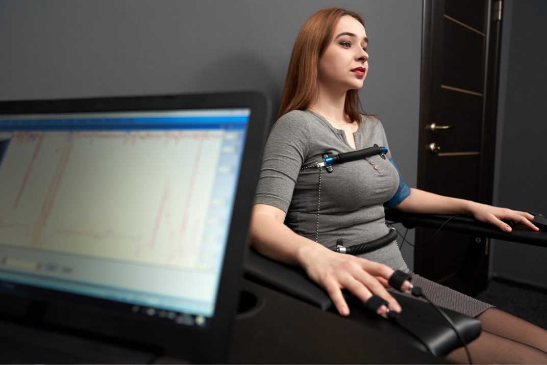 Woman sitting, connected to a polygraph machine, with laptop displaying data, in a room.