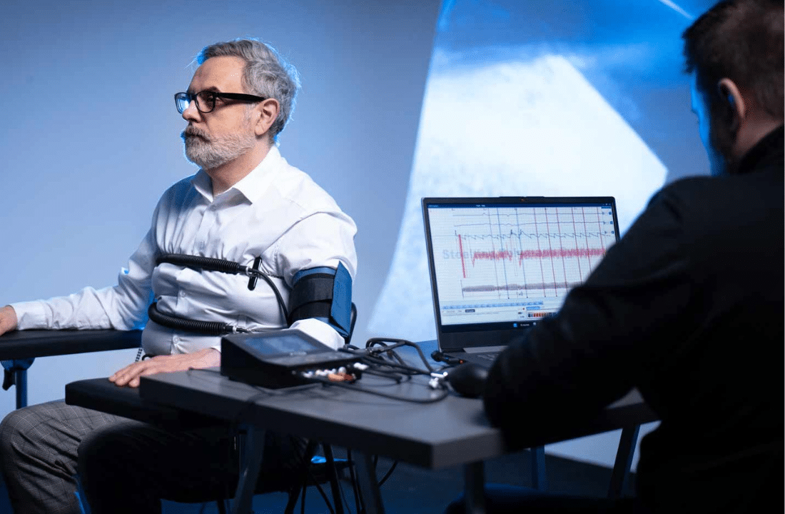 Man in a polygraph test, connected to monitoring devices. A person is seated at a table with a laptop.